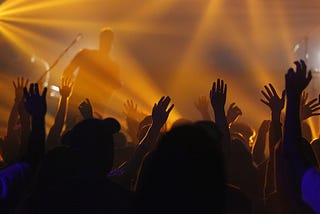 A crowd seen from behind while they cheer a band submerged in orange lights.