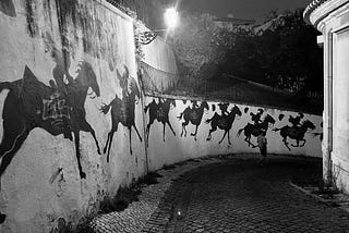 A nighttime view of a street in Lisbon, the cobbled calçadas gleaming in teh streetlight, the wall painted with a horse and rider galloping further and further away around the bend.