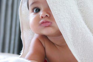 A colour photo of a pouting brown-eyed baby with a white towel partly over its head.