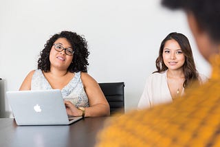 Two individuals sit across a table from a third person. There is a laptop in front of one of the two on the far side of the table.