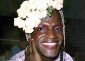Portrait of Marsha P. Johnson smiling with white flowers in her hair and mauve makeup