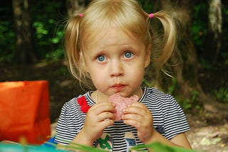 A young girl looking as as she holds a slice of salami in her hands, close to her mouth