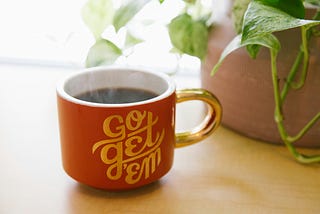A coffee cup sits on a desk in front of a potted house plant. The side of the cup reads ‘Go get ‘em’.