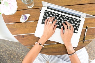 Computer on a planked desk and female hands with rings and bracelets resting on the keys.