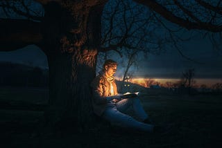 A man sitting beneath a tree at night, reading a book. The book shines a golden light.