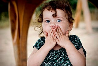 A white girl about age four with light brown curly hair holds her hands to her face in a surprised expression. She has big blue eyes and wears a green and white polka dot T-shirt. She is in focus with a few wooden structures out of focus in the background.