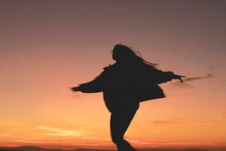 The silhouette of a person with long hair and glasses dancing on a sunset beach.