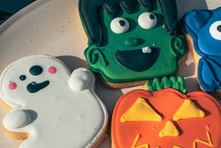Decorated Halloween cookies shaped like a ghost, Frankenstein’s monster, and a jack-o’-lantern on a white plate — symbolizing how fear is sweetened and made harmless.