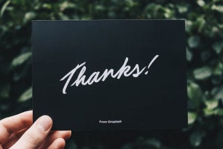 Left hand holding a black card that says “Thanks!” in a cursive font, and then smaller below says “From Unsplash.” Card is held against a green leafy background