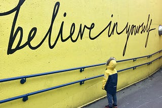 A young child looks up at a yellow wall with “believe in yourself” in cursive