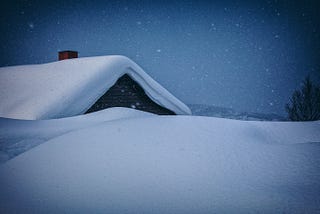 A nighttime shot of a snowy rooftop with a square, brick chimney peeking up above the blanket of snow.
