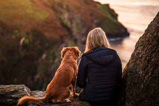 A woman and her dog sit on a rock and look out over the water together