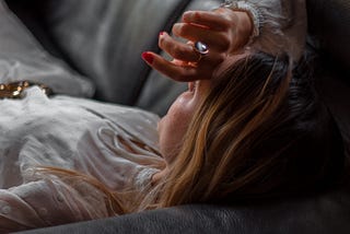 Woman dressed in white satin lies on a couch with her arm over her forehead. She has an opal ring on her middle finger, light brown long hair, white skin. We only see the side of her face, her hand and her chest.