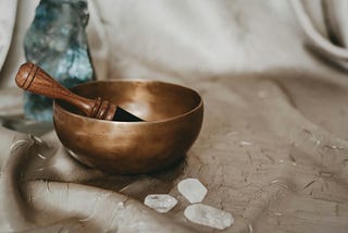 A draped white cloth with, a meditation bowl bell with hammer, crystals, and scattered filaments sitting on it.