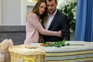 A man and a woman at a funeral, looking at a straw coffin