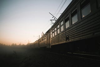 Photo of a passenger train heading off into a foggy horizon. The colors are drab and the image is gloomy and mysterious. The train might be derelict. Photo by Andrey Svistunov on Unsplash.