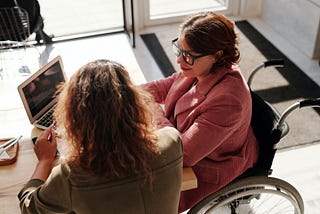 A women in a wheelchair looking at a computer screen with another women sitting next to her.