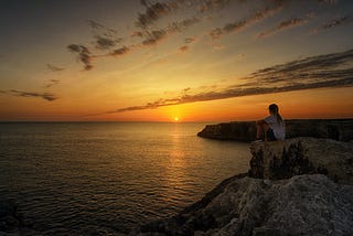 person sitting on a cliff looking at still water during a beautiful sunset