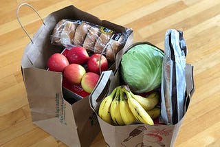 Two brown shopping bags filled with food such as bananas, red apples, bagels, iceberg lettuce and bread.