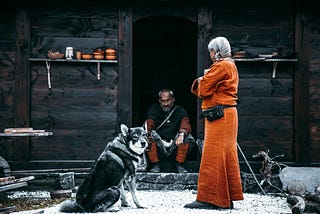 A viking women dressed in all orange stands in front of a Viking man dressed similarly along side a dog