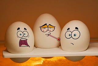Three white eggs with cartoon faces expressing different emotions — one frightened, one sick with a thermometer, and one worried — sitting together on a shelf under warm lighting, symbolizing fear, anxiety, and vulnerability in a humorous way.