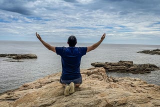 A person kneels on the beach, their back to the viewer, facing the ocean, with their arms raised to the sky.