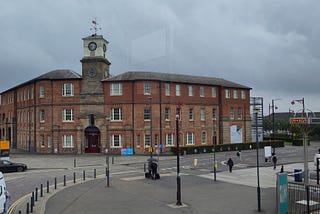 View across a wide pavement and road towards an old brick three-storey building on a corner. A stone clocktower that is taller than the rest of the building is set at the corner, which wings in brick either side