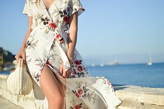 Girl wearing cream dress with floral print, carrying a sun hat by the sea