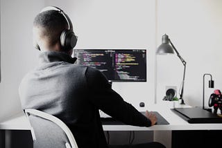 Man in black long sleeve shirt wearing black headphones, sitting on a chair, looking at a computer screen with code editor open