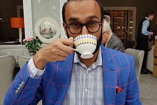 Brown man with tortoiseshell glasses and beard with a teacup raised to mouth in right hand. Wearing a light blue blazer with a red windowpane check and a blue check shirt underneath.