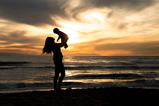 A mother holds up her young child upon a beach. The ocean is calm. The clouds swirl in the sunset.