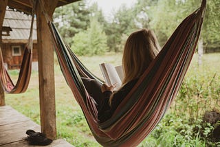 Woman reading a book in a hammock that’s strung up on the porch of a rustic cabin with grasses and flowers beside it.