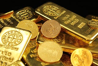 The image shows a collection of gold bullion bars and coins, gleaming with a bright yellow hue indicative of high purity. The bars are stamped with markings that denote their weight (1 kilo), purity (Fine Gold 999.9), and the name of the refinery (likely “Argor-Heraeus SA” based on the visible text, which is a well-known precious metals refiner based in Switzerland). The coins feature various designs, with some showing an animal, possibly a kangaroo, which suggests they might be Australian Gold.