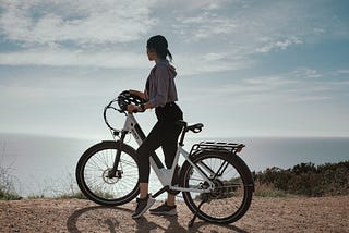 A young woman straddling an ebike, standing on the ground, and looking out over a body of water