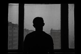 The silhouette of a man looking out of a window at several plain concrete buildings. The sky is overcast and everything is gray.