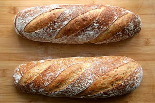 This is a photo of two loaves of unsliced bread. They may be homemade or from a bakery and they have flour visible on top.
