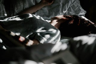 A person lies on white sheets, surrounded by warm sunlight patches. The face is turned away.