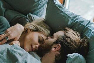 Blonde woman being kissed on the forehead by brown haired man while they lay in a bed with grey sheets