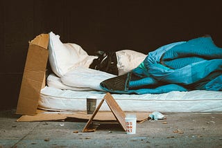 A makeshift bed on a concrete floor, comprising a white mattress and pillow, a blue blanket and large pieces of cardboard.