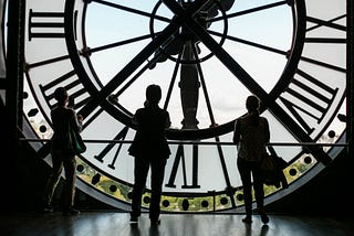 giant wall clock with 3 figures looking up to signify TIME