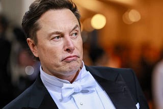 a picture of elon musk doing that weird thing he does with his lip where he like, bunches it up. he’s also raising his eyebrow in a way that makes him look like a character from a 2000s DreamWorks movie. oh, and he’s wearing a suit with a white bow-tie. the background is blurry, as if you are drunkenly focusing on the most annoying man in the room.