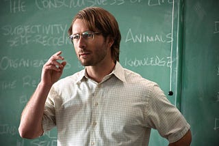Gary, the man character, standing in front of a chalkboard as he teaches class. He is wearing a short-sleeved button-down shirt and glasses. His hair is a weird length that most would not consider fashionable (they are wrong).