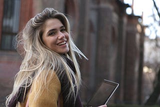 Confident blond, smiling woman looking back at us over her shoulder while holding an i-Pad. She’s outside and wearing a thick wooly jumper