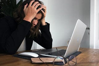 Image of woman in distress, gripping her head with her hands while staring at a laptop. Her glasses and phone are in the foreground next to her computer