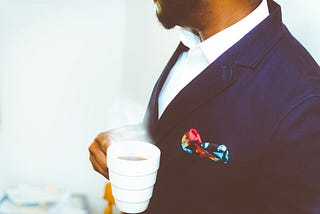 Black man with a beard in an expensive dark blue suit and crisp white shirt holding a cup of coffee.