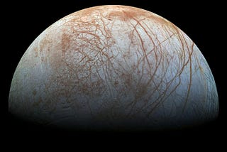 The surface of Europa against the blackness of space. The surface is light blue and white with reddish cracking all across it.