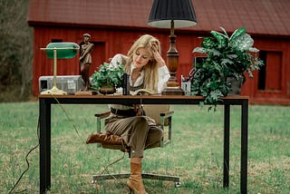 Brown table, with 2 lamps, printer 2 flower plants on it. I woman sitting at the table with her head resting on left hand. The table looks like an office table. She is outside. You can see a red barn in the distance.