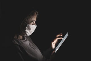 A woman wearing a mark reads a tablet in a dark room