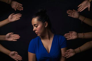 A woman lying down with her eyes closed, her head tilted to her right, on a surface surrounded by the hands of other people.