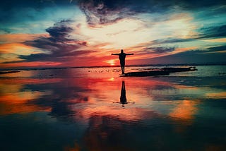 This is a picture of a brilliant sunset. A silhouette stands on the beach where the waves meet the shore, arms outstretched in the form of a cross, giving the image a spiritual feel.